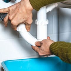 Close-up of a person fixing plumbing under a sink. They are using both hands to tighten or loosen a white plastic pipe joint. A blue plastic bin is placed underneath the pipes, likely to catch water or debris.