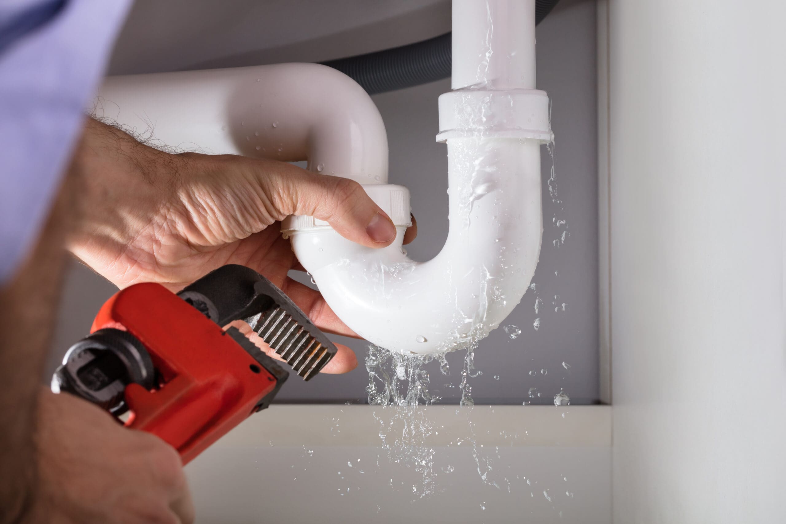 A person uses a red pipe wrench to tighten a leaking white plastic U-bend pipe under a sink. Water is dripping from the pipe as it's being adjusted. The pipe is part of a plumbing system beneath a countertop.