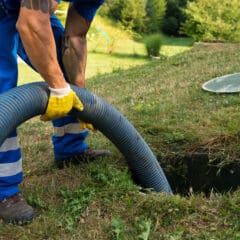 A worker wearing blue overalls and yellow gloves is holding a large hose, inserting it into an open septic tank in a grassy outdoor area. Nearby, the manhole cover is placed on the grass.