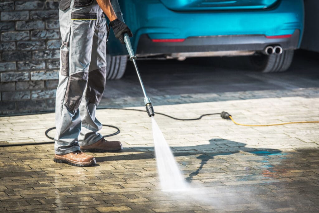 A person wearing gloves and protective clothing uses a pressure washer to clean a paved driveway. A blue car is parked in the background. The foreground shows streams of water spraying over the ground, cleaning the surface.