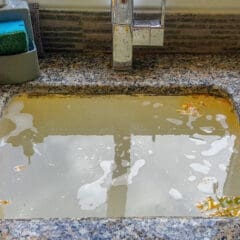 A kitchen sink filled with dirty, soapy water and food debris. The water is murky, with food particles floating on the surface. A sponge and scrub brush are in a holder next to the sink. The countertop has a granite finish.