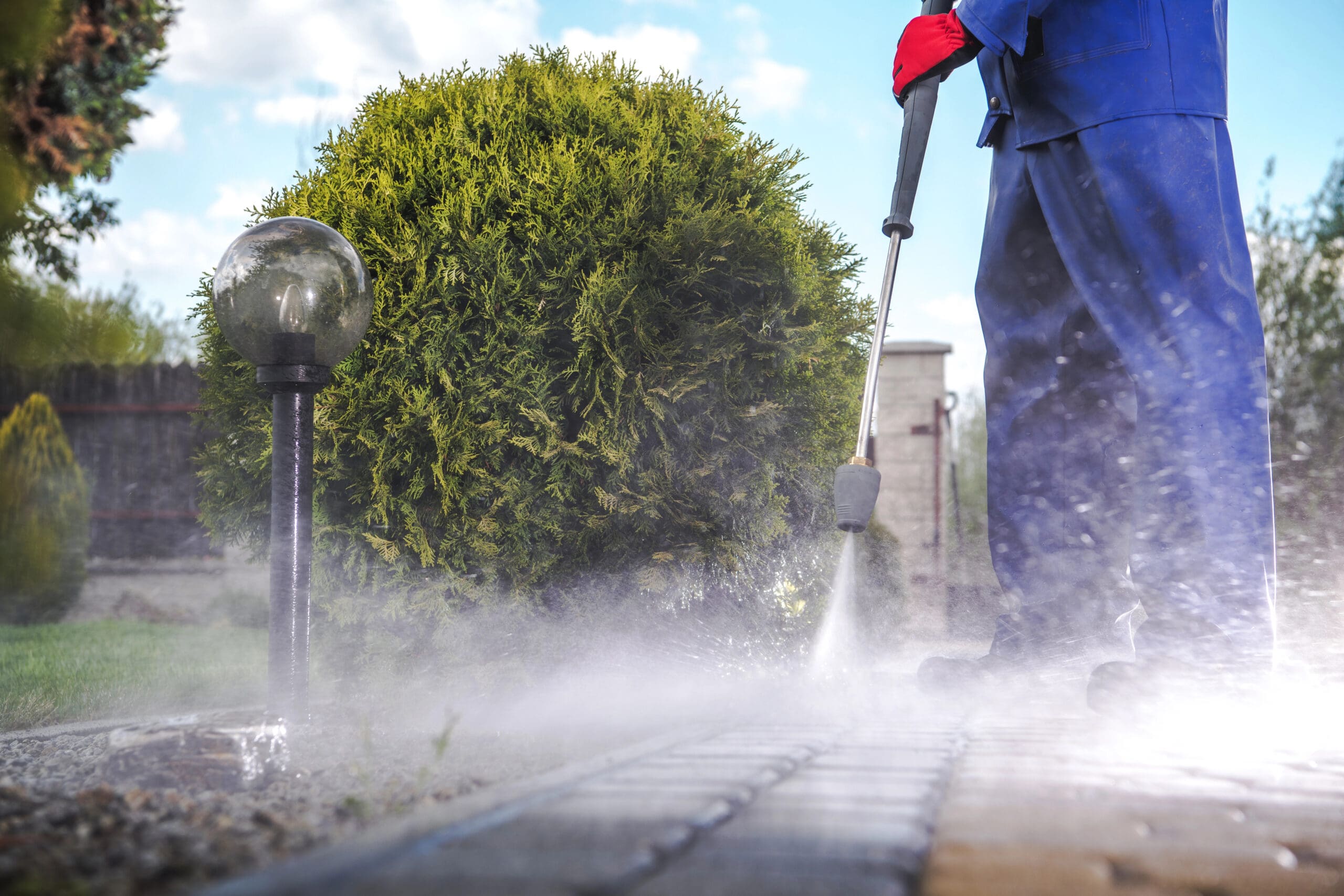 A person dressed in blue overalls and red gloves is pressure washing a paved walkway in a garden. A small tree and an outdoor lamp are visible in the background, surrounded by grass and greenery.