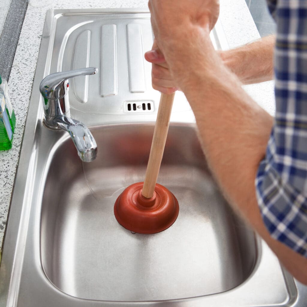 A person wearing a blue plaid shirt is using a plunger to unclog a stainless steel kitchen sink. The sink area is clean, with a bottle of dish soap visible on the left side. The faucet is positioned over the sink's basin.