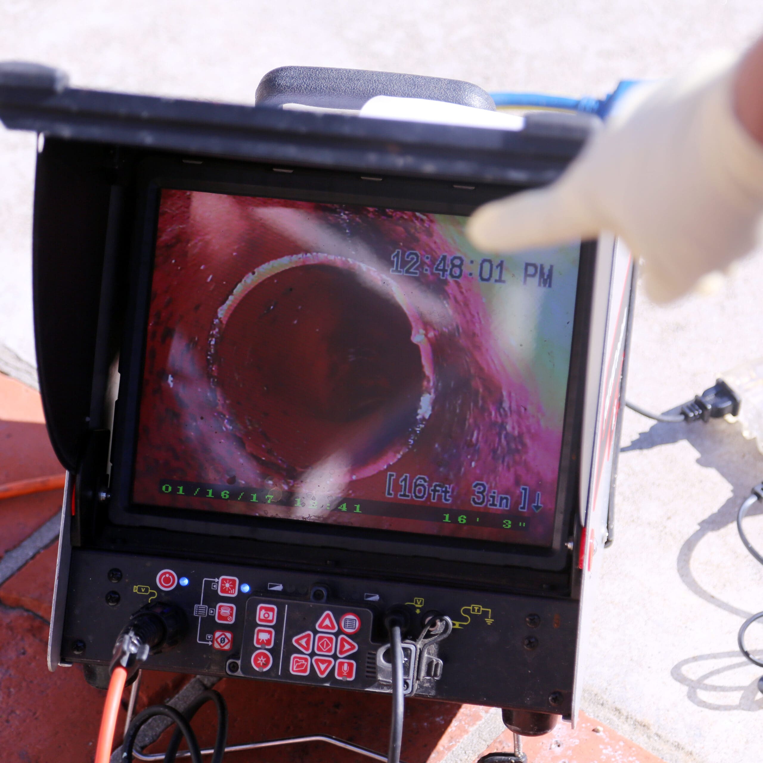 A gloved hand points at a screen displaying an image from a sewer camera inspection. The screen shows the interior of a pipe, with the distance measurement of 16 feet 3 inches displayed. Various cables and control buttons are visible around the monitor.