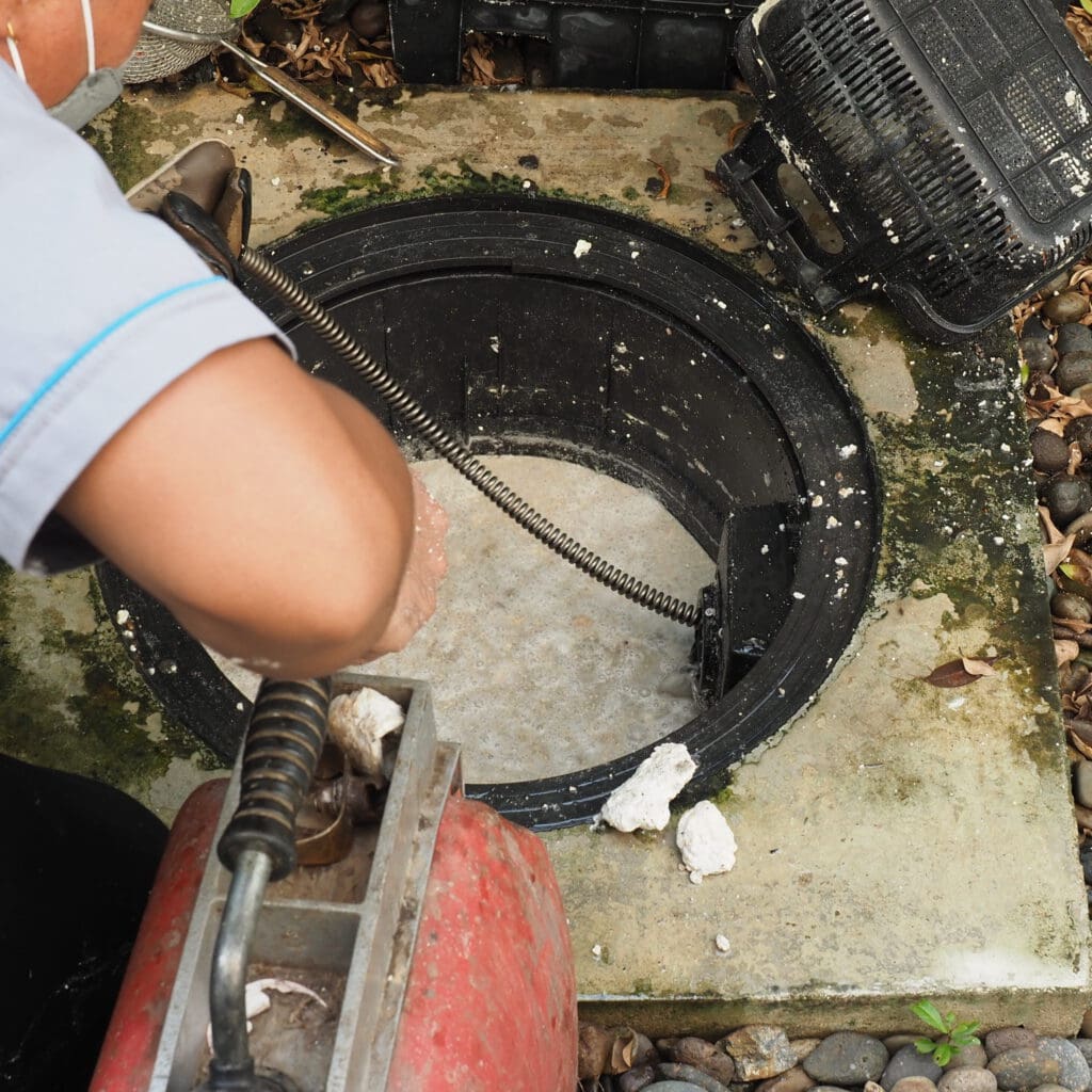 A person cleaning a manhole with a mechanical drain cleaner. The person’s hands and part of their arm are visible as they operate the equipment, which is being used to clear debris and buildup in the manhole. The surrounding area has leaves and a black plastic container.