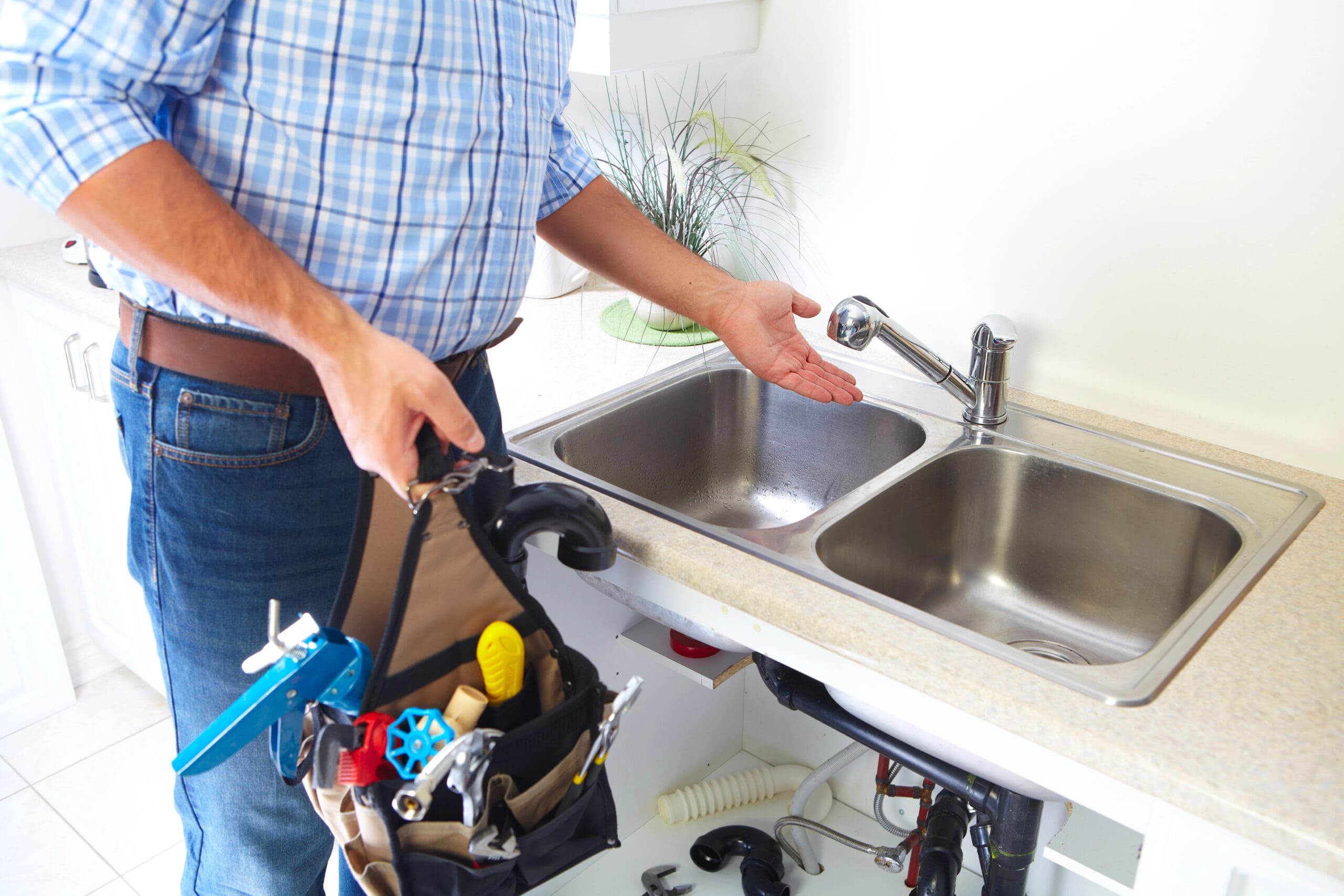 A person in a plaid shirt and jeans stands near a kitchen sink, holding a tool belt filled with various tools and plumbing supplies. The sink area is clean and modern, and the person is gesturing towards the faucet.