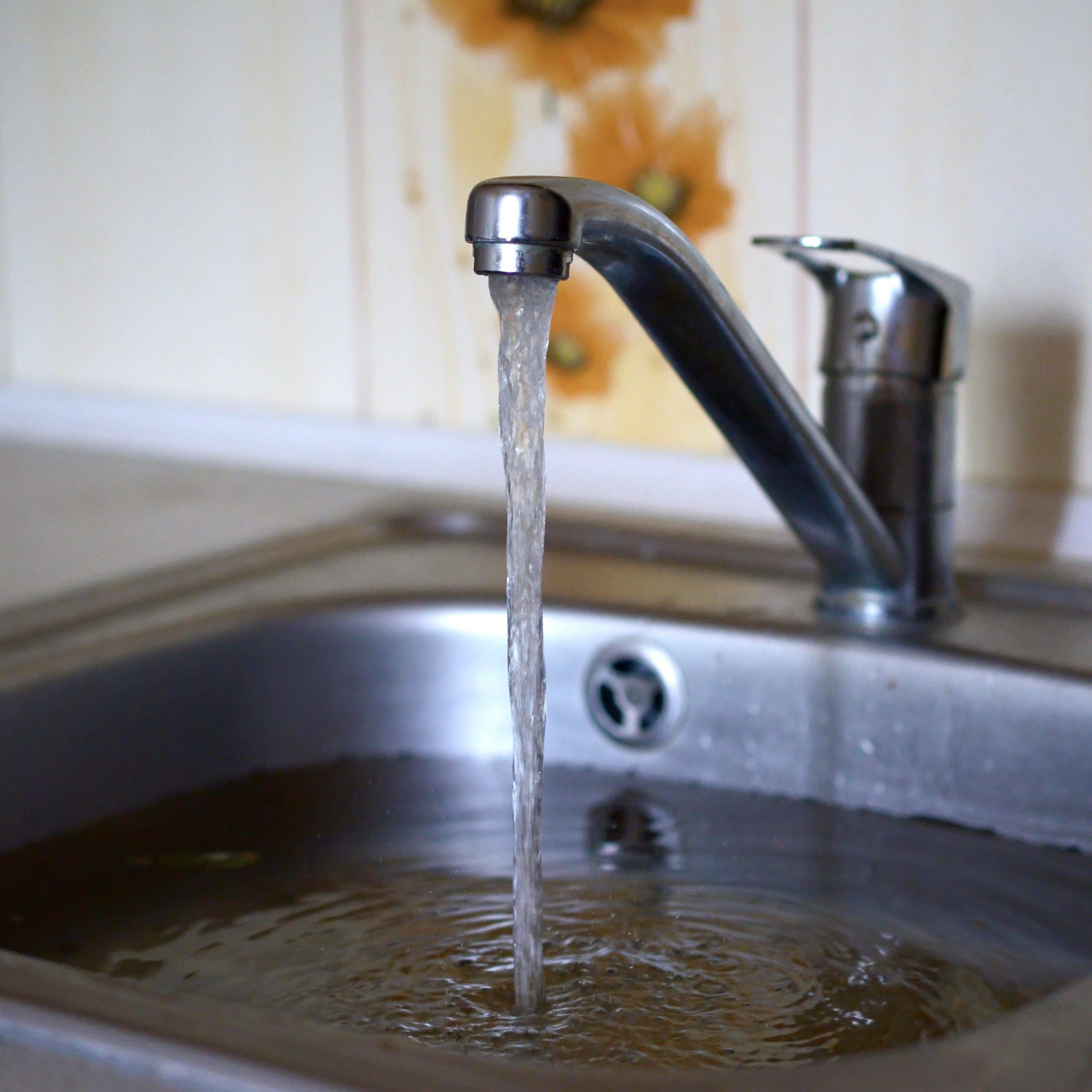 A silver kitchen faucet with water running into a stainless steel sink. The water appears to be discolored, suggesting possible contamination. The background includes a light-colored wall with a sunflower design.