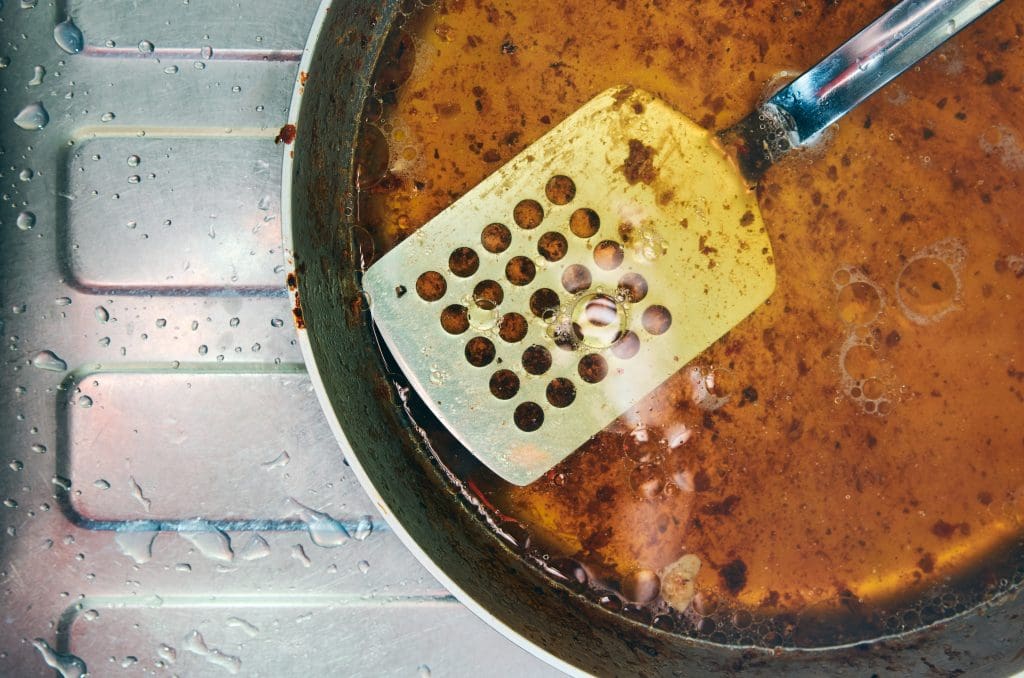 A dirty frying pan filled with greasy, brown oil and food residue sits in a metal sink. A metal spatula with perforated holes rests inside the pan, coated with the same greasy residue. Water droplets are visible on the sink surface.