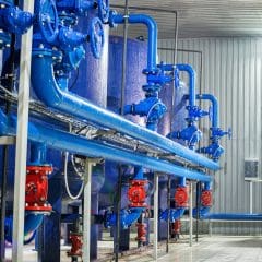 A brightly lit industrial facility with blue pipes and machinery against a metallic wall. The pipes are interconnected and some are fitted with red valves and connectors. The floor is clean and reflective. An air conditioner is visible on the wall.