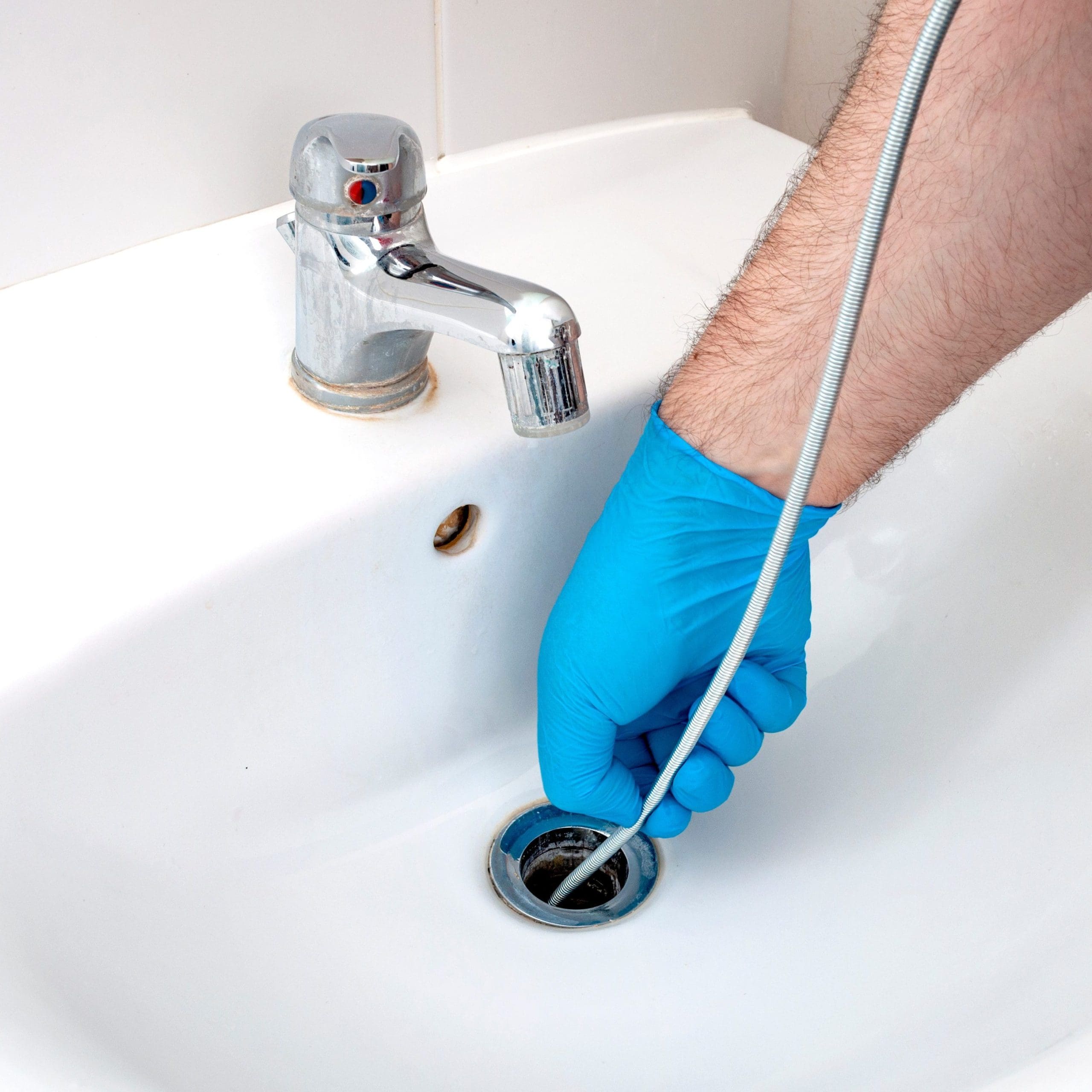 A person wearing blue gloves uses a drain snake to clear a clogged bathroom sink. The sink features a chrome faucet, and the action is focused on the drain area.