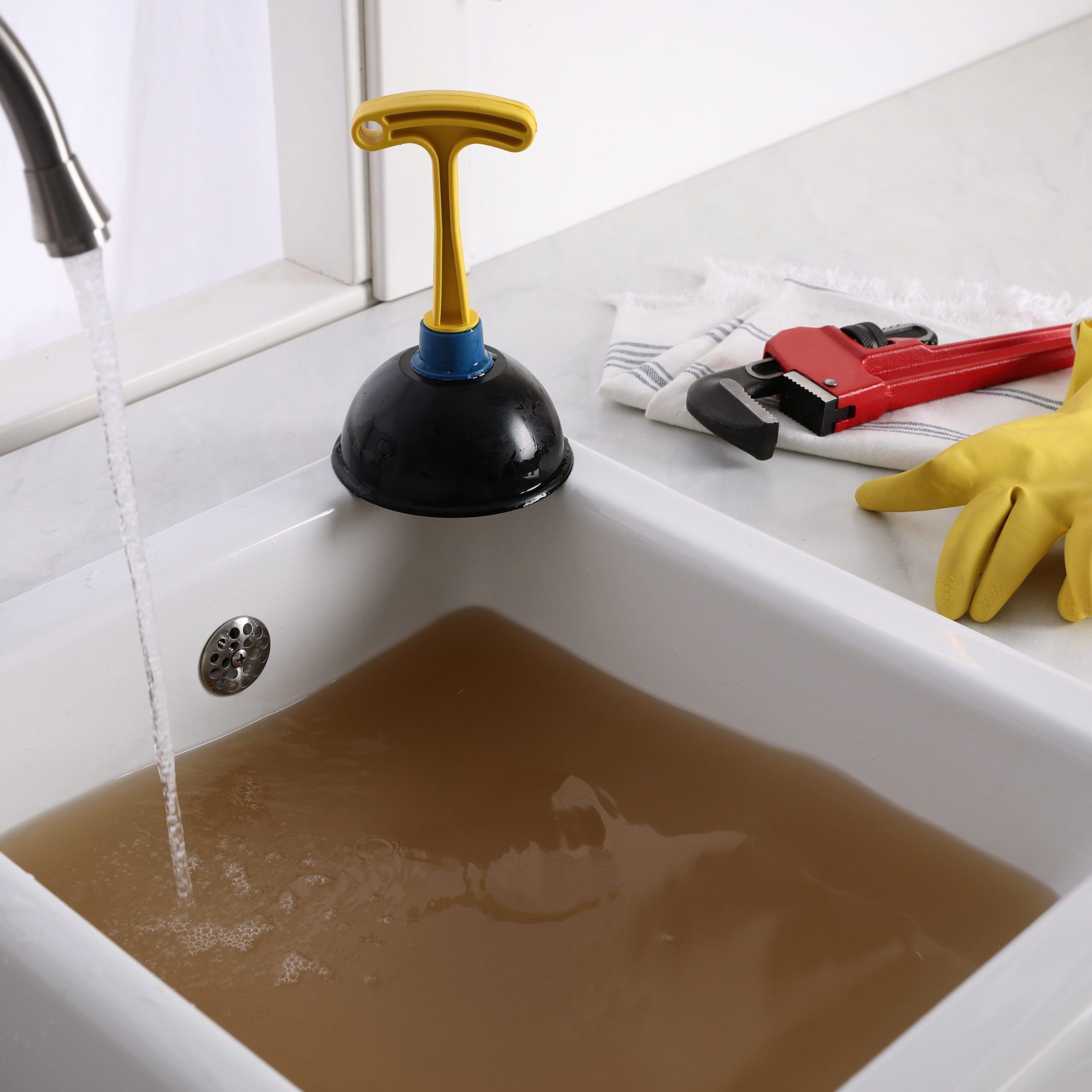 A white sink filled with dirty brown water. A plunger is placed upside down on the sink’s edge, and the faucet is running with clear water. Nearby are a pair of yellow rubber gloves, a red wrench, and a white towel, all on the countertop.