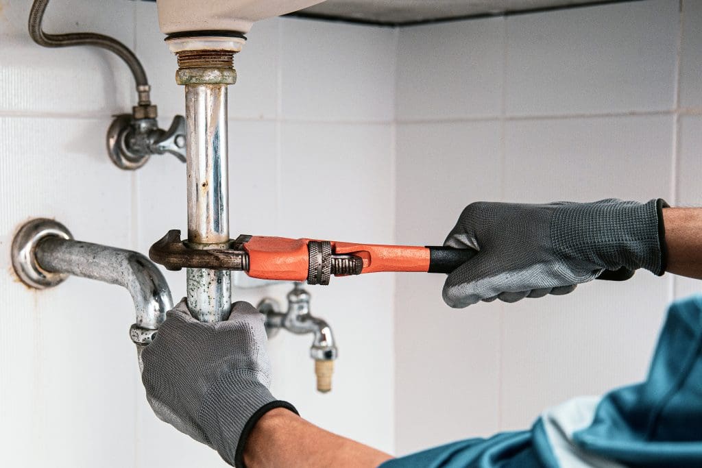 A plumber wearing gray gloves uses an orange pipe wrench to fix a pipe under a sink in a tiled bathroom.