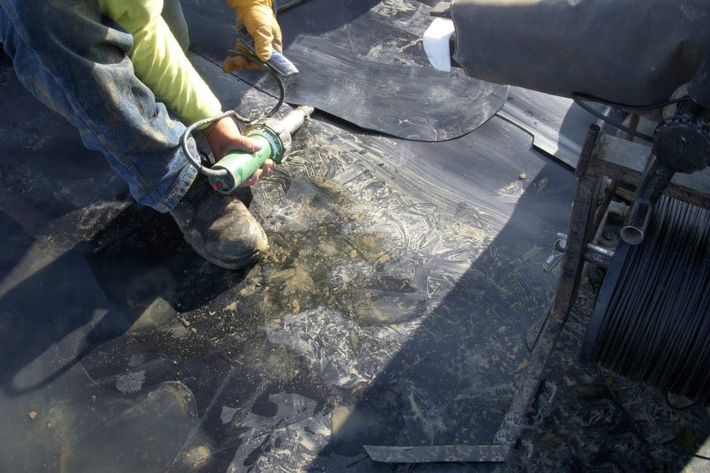 A construction worker is using a heat gun to weld a seam on a black rubber roof membrane. The worker's hands are wearing yellow gloves, and the person is wearing blue jeans. Various construction materials and tools are visible around the work area.