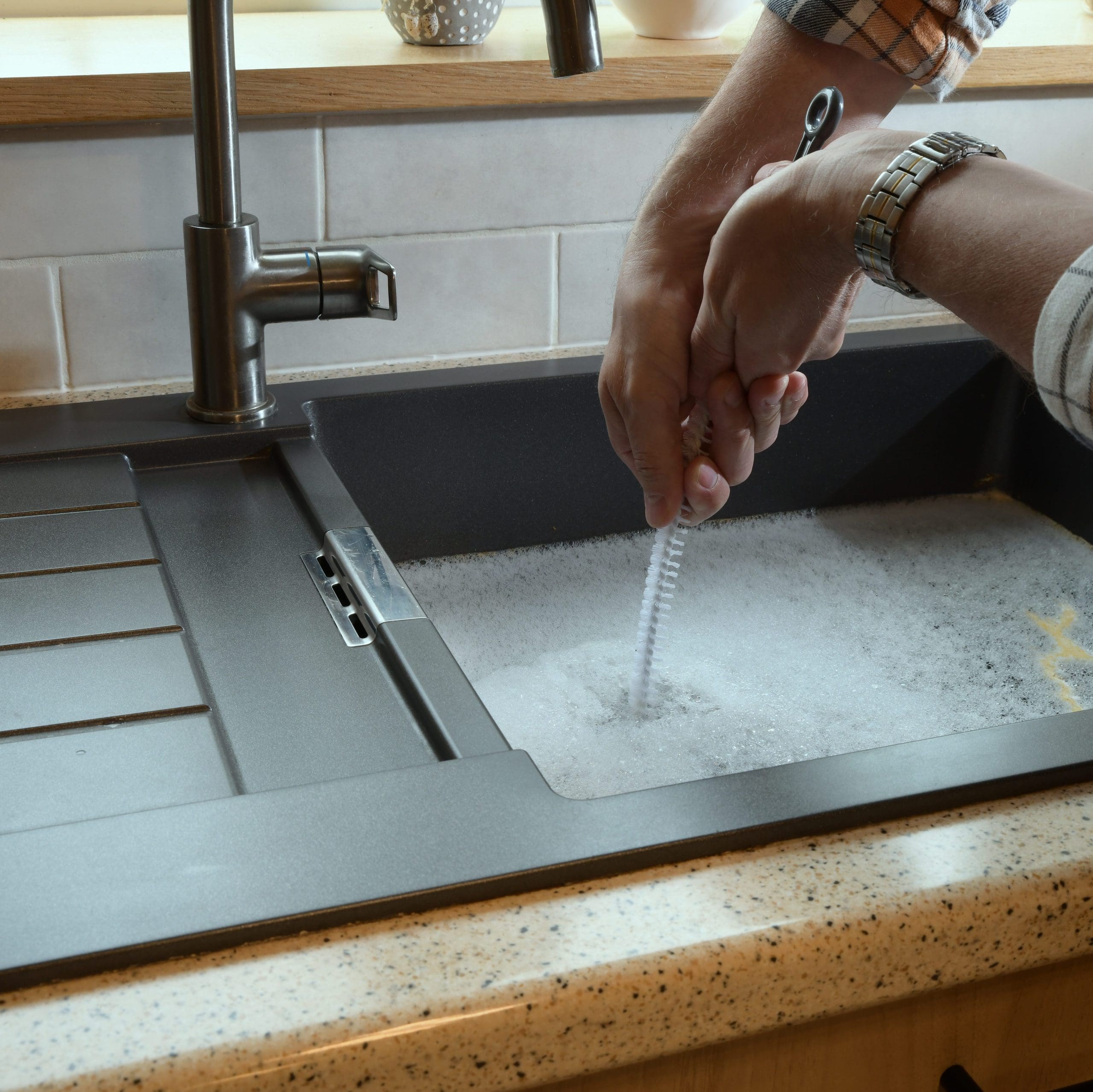 A person is washing a small brush in a kitchen sink filled with soapy water. The sink area features a modern faucet, a black drying rack, and beige countertops. The person is wearing a plaid shirt and a wristwatch.