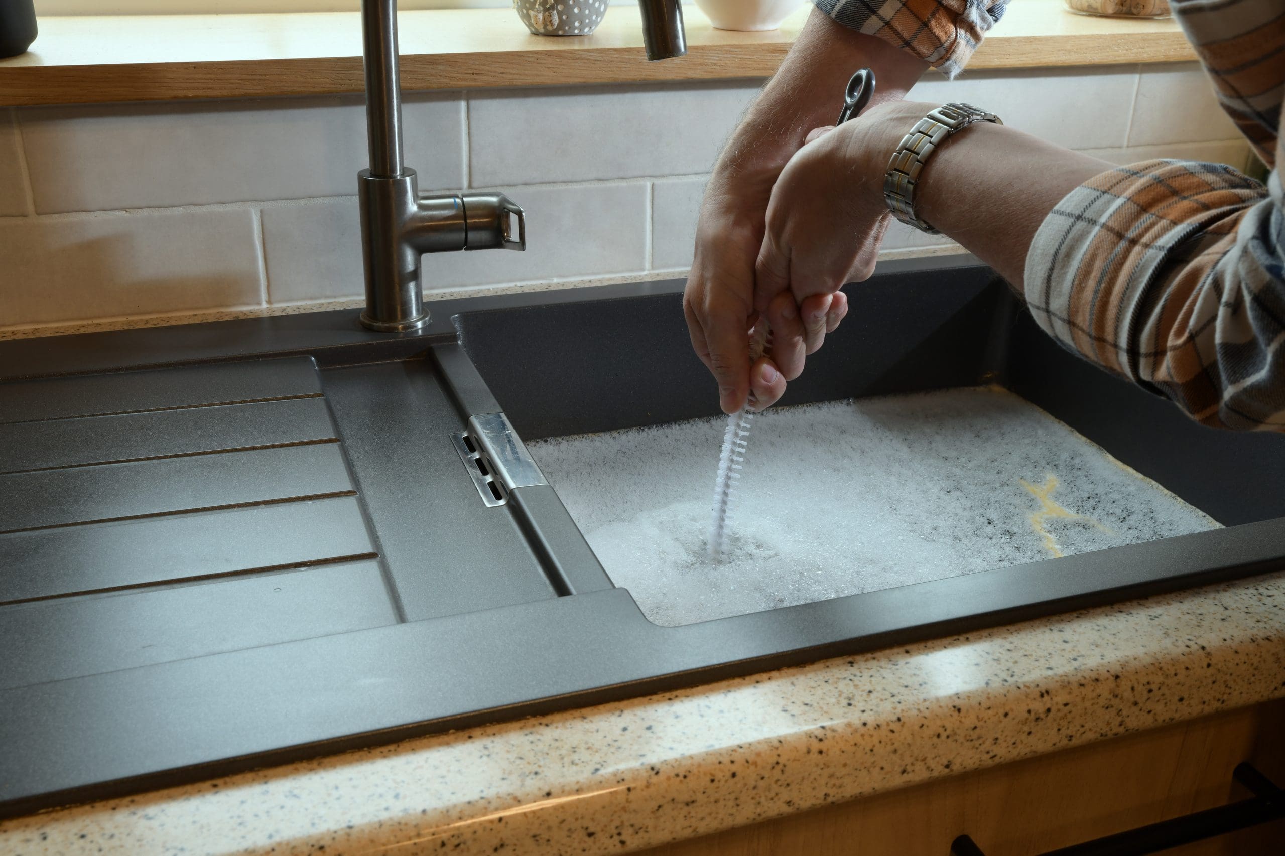 A person in a plaid shirt is cleaning a dish with a soapy brush in a sink. The sink area is tidy, with a modern tap and a textured countertop. The surroundings include a backsplash with light-colored tiles and some decor items on the windowsill.