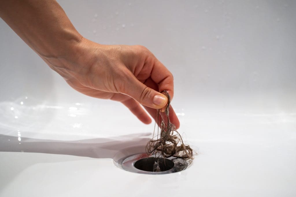 A hand is seen pulling a clump of hair from a white sink drain. The background is blurred and the focus is on the hand and the hair. The sink is clean and the drain is in the center of the image.