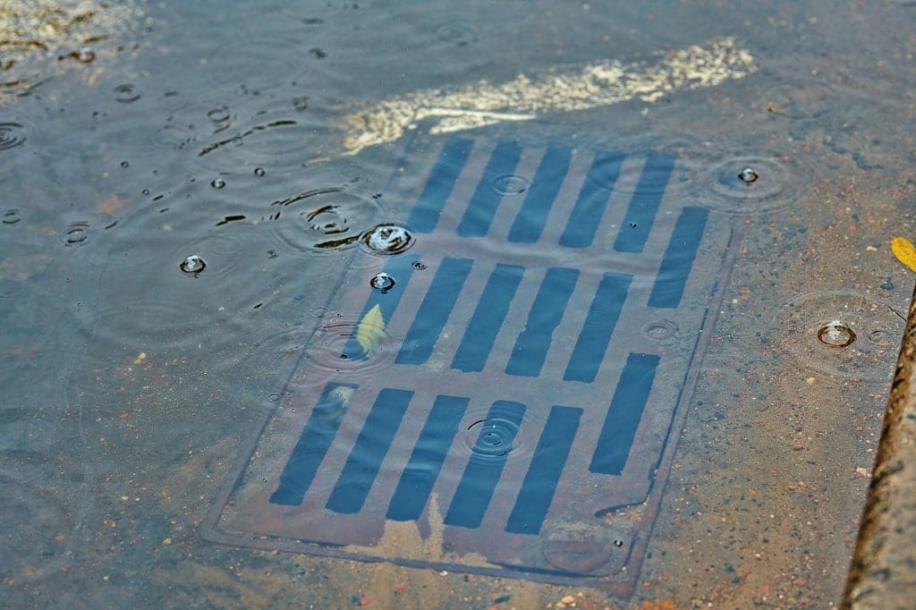 A storm drain partially submerged in rainwater on a paved surface, with raindrops creating ripples on the water's surface. The pavement has some scattered debris and a small patch of wet leaves nearby.