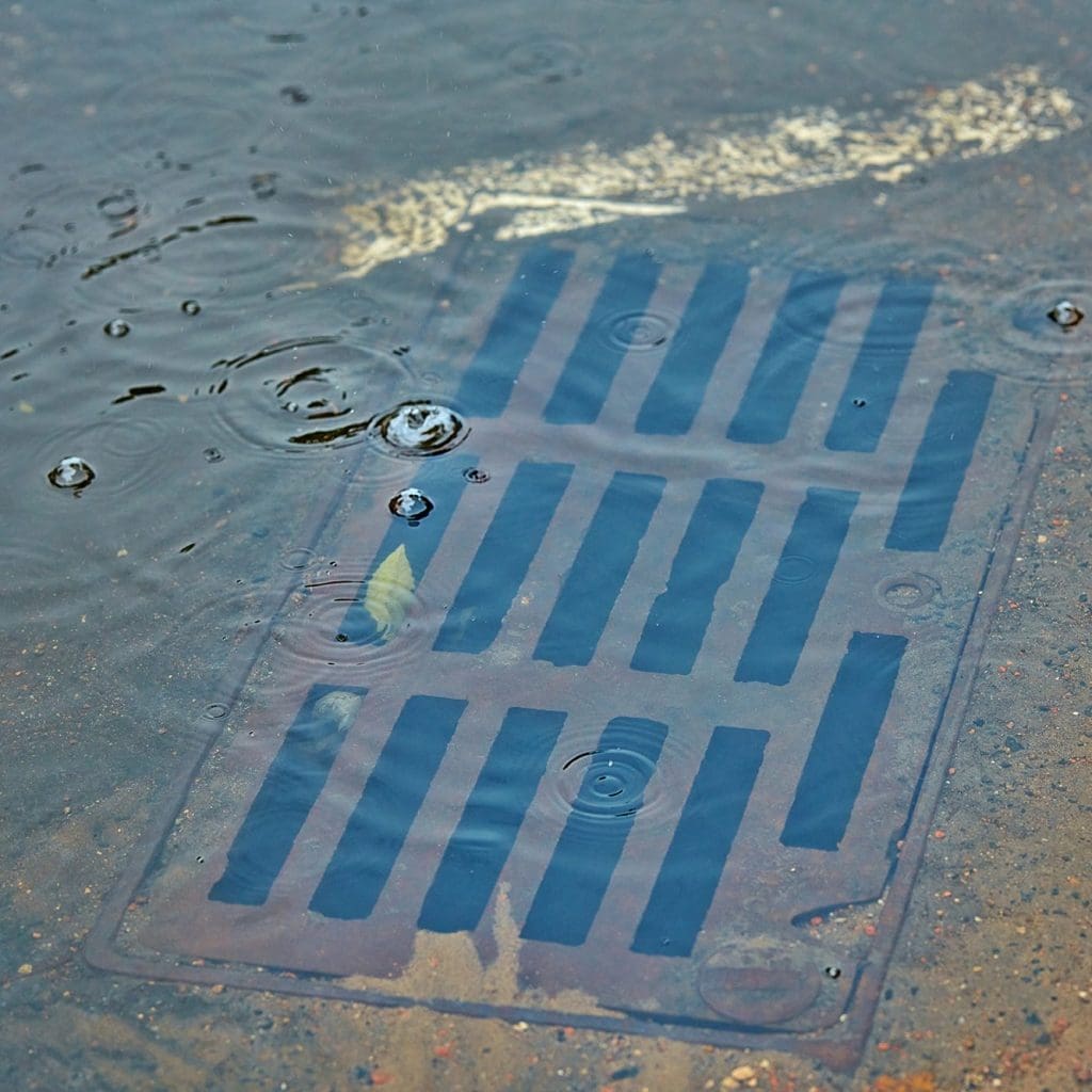 A storm drain partially submerged in water with raindrops creating ripples on the surface. The steel grate has vertical and horizontal slots and is surrounded by a wet concrete surface. A faded white mark is visible near the top of the drain.