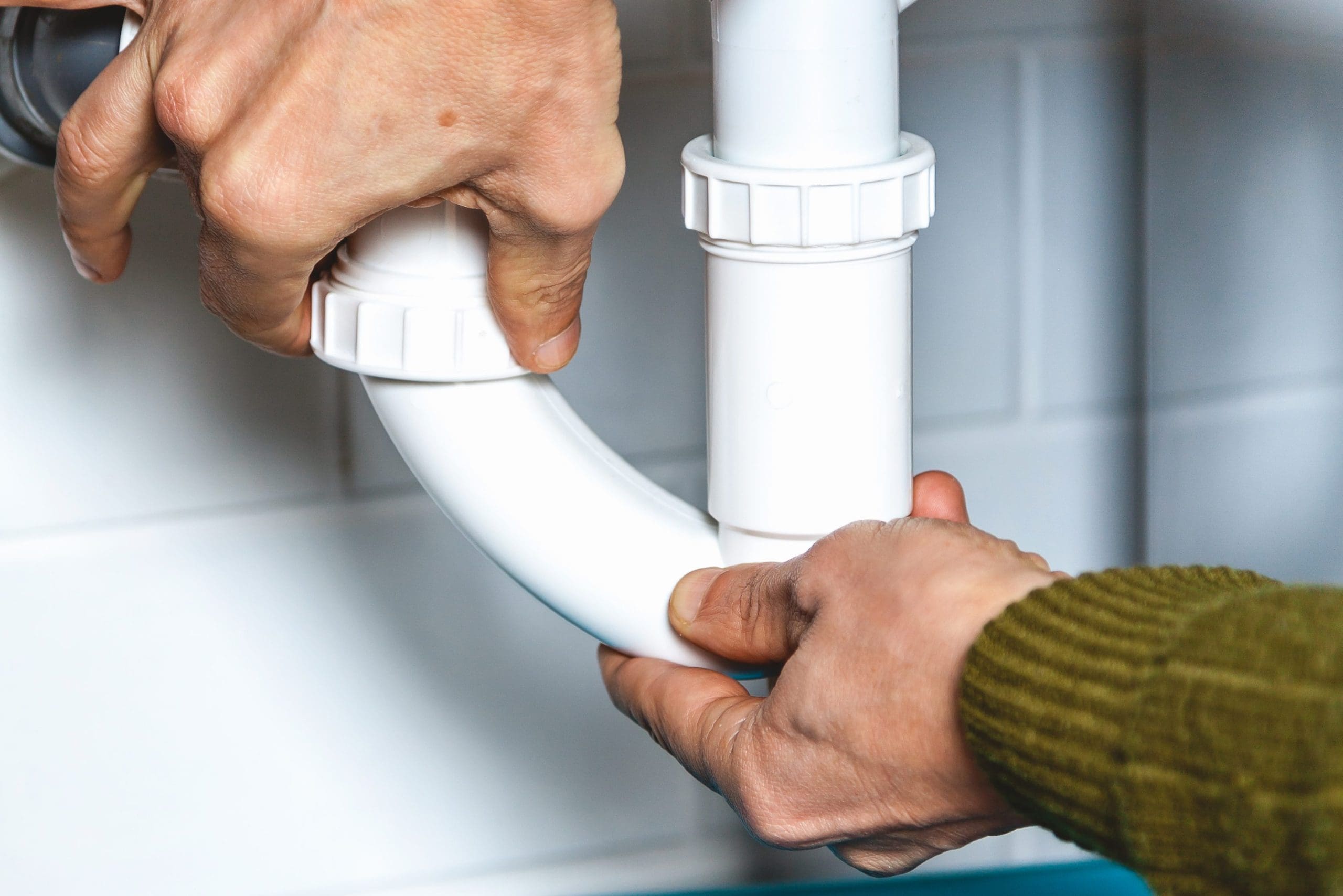 Close-up of two hands fixing a white plumbing pipe under a sink. The person is tightening a connector on the curved part of the pipe, ensuring a secure fit. The background features white tiled walls.