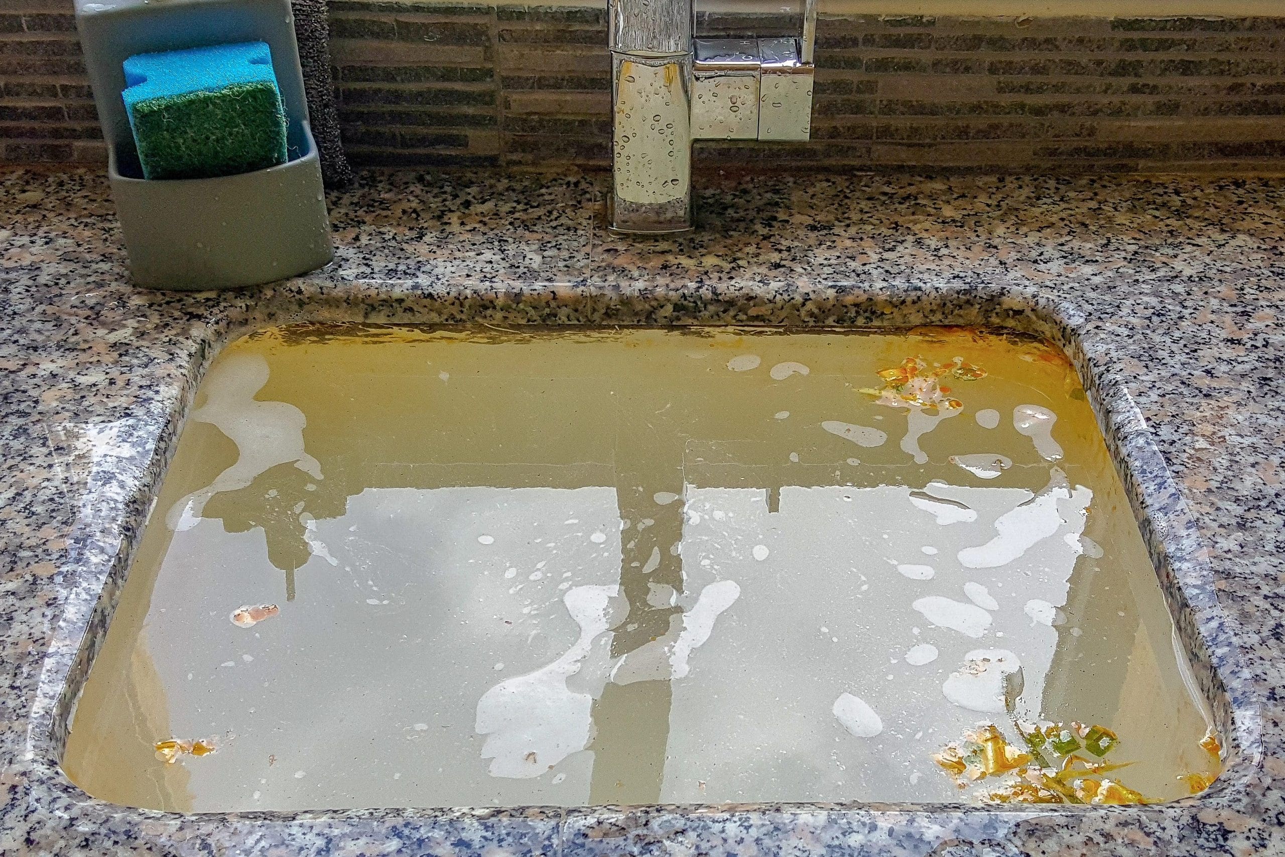 A kitchen sink filled with cloudy, dirty water and some food particles floating on the surface. Above the sink is a faucet with water droplets, and next to it is a sponge in a holder. The countertop is speckled gray, and the backsplash is tiled.