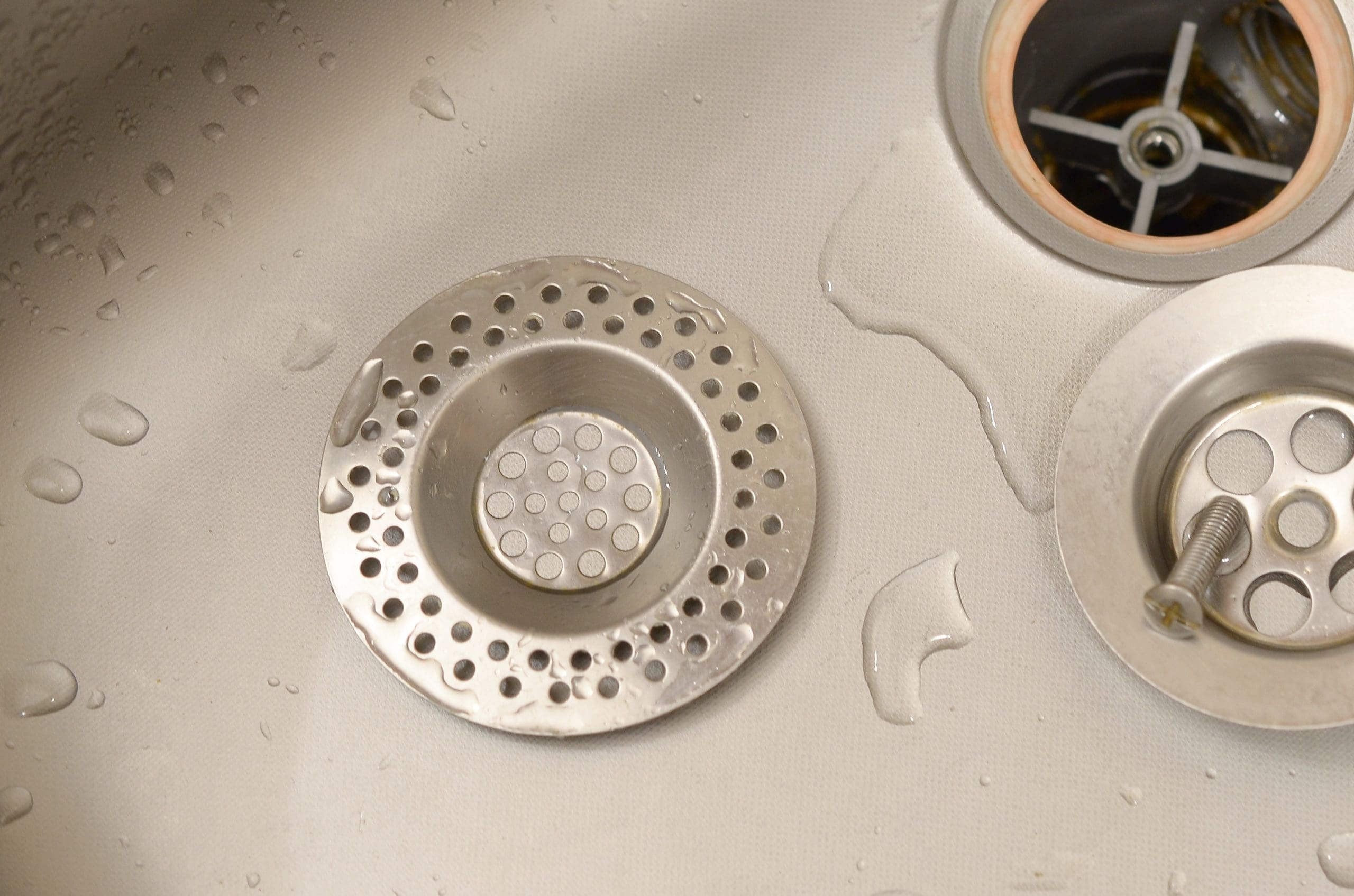 A close-up image of a metal kitchen sink showing a removed round sink drain strainer with multiple holes. The strainer is placed next to the drain opening, with some water droplets visible around it on the sink surface.