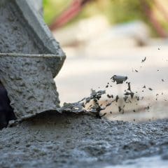 Close-up of wet concrete being poured from a chute onto the ground. The concrete splashes as it hits the surface, creating a textured and dynamic scene. The background is slightly out of focus, emphasizing the motion of the pouring concrete.