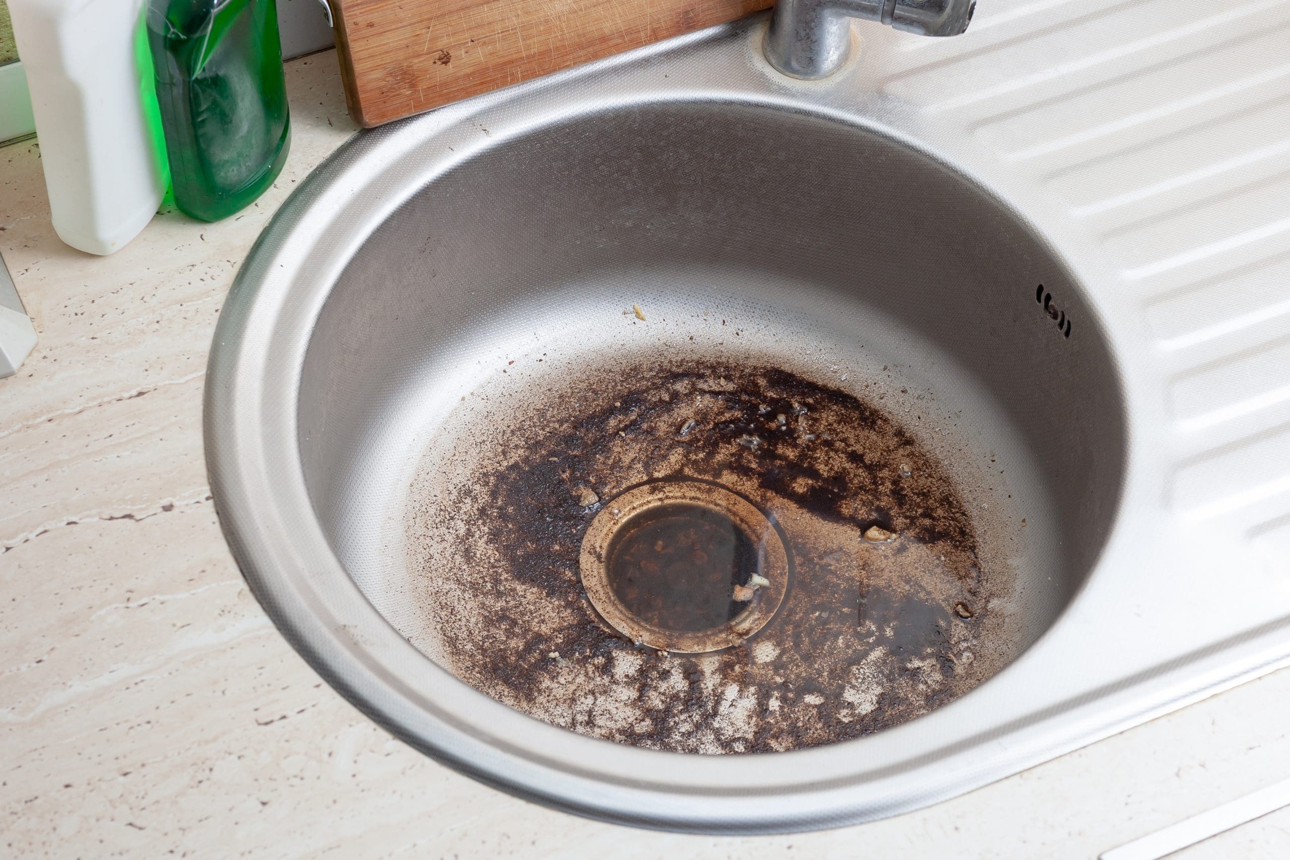 A metal kitchen sink with a dirty basin showing brown stains and grime near the drain. A green spray bottle and a white cleaning product sit to the left on the countertop, next to a wooden cutting board. The faucet is partially visible, attached to a drainboard.