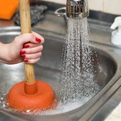 A person's hand with red-painted nails holds a wooden-handled plunger in a stainless steel sink full of water. Water is running from a faucet into the sink, splashing around the plunger. Soap, a scrub pad, and other kitchen items are visible in the background.