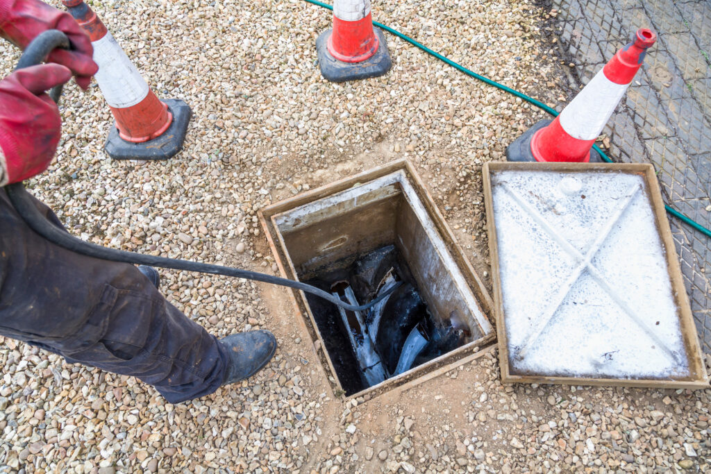 A worker wearing gloves stands next to an open utility manhole with cables inside, surrounded by traffic cones on a gravel surface. The manhole cover is set aside.