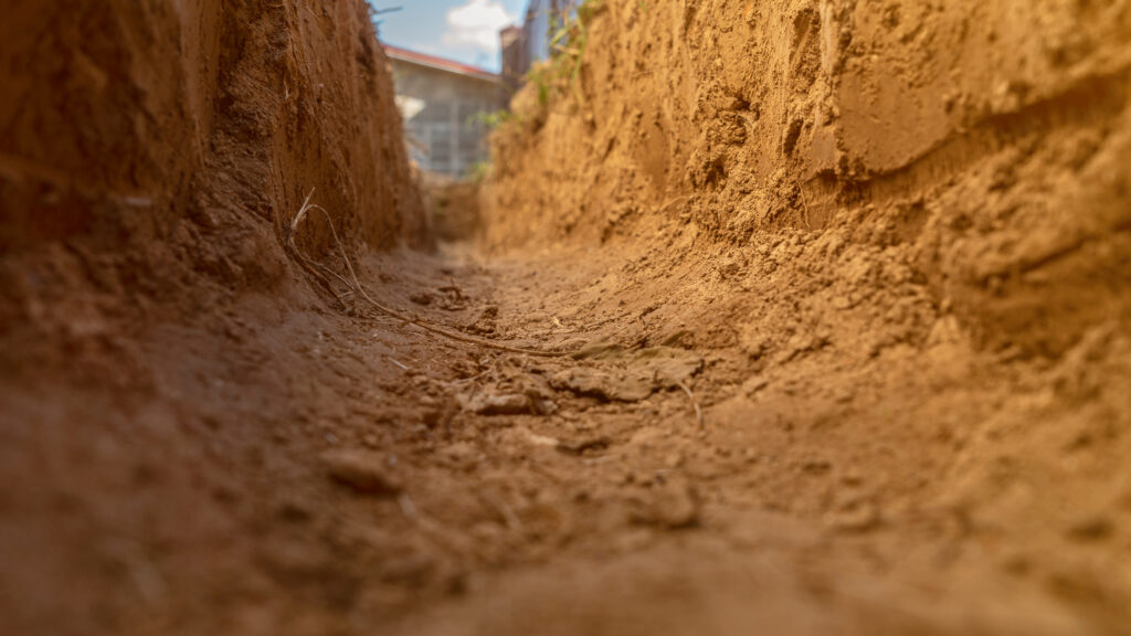 A close-up view from inside a shallow dirt trench, with textured soil walls and a dry, rocky ground. Sunlight illuminates the scene, and a blurry building is visible in the background.