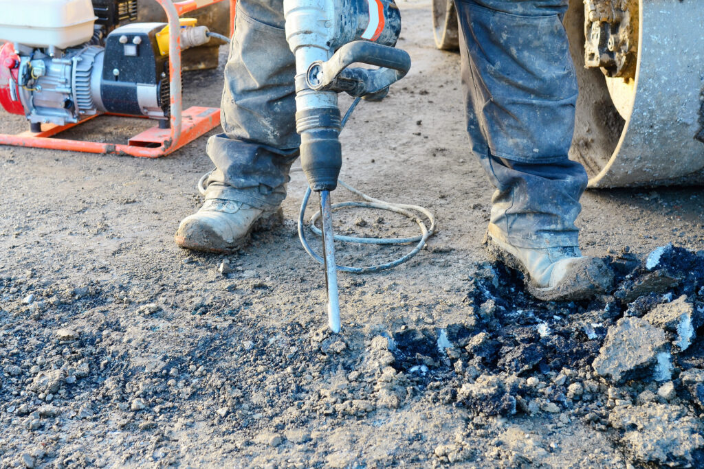 Worker in construction boots uses a jackhammer to break up asphalt on a road. Debris and a roller machine are visible nearby, with loose pieces of asphalt scattered on the ground.