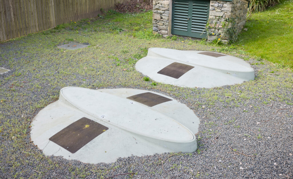 Two concrete covers with metal plates embedded in grassy ground, possibly for underground utilities or tanks, are situated near a stone structure with a vented green door and a wooden fence in the background.